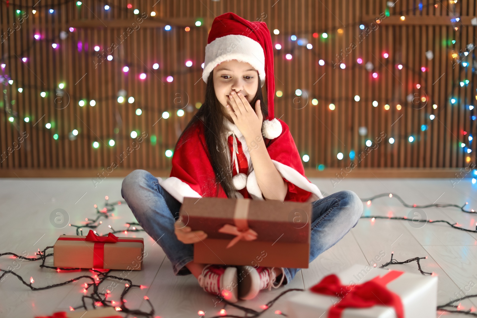 Photo of Cute little child in Santa hat opening Christmas gift on blurred lights background