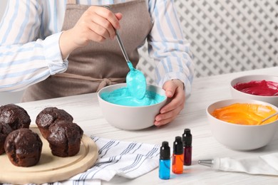 Photo of Woman mixing cream with light blue food coloring at white wooden table, closeup. Decorate cupcakes