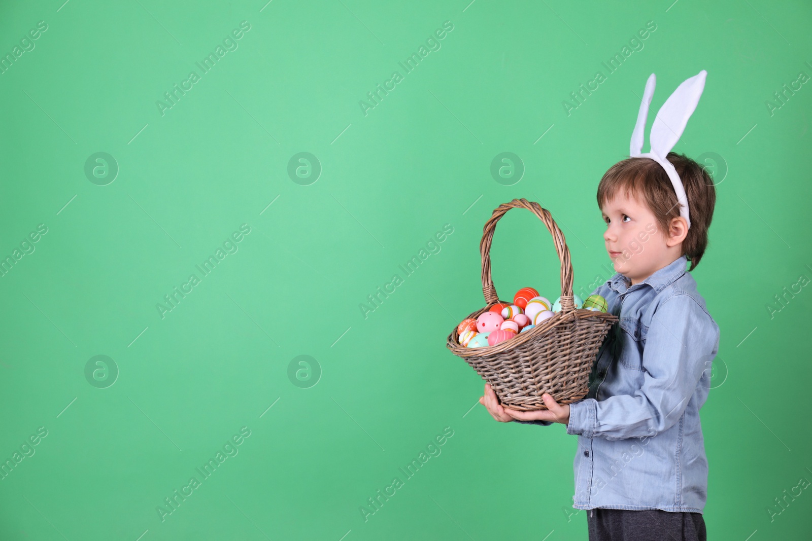 Photo of Cute little boy wearing bunny ears with basket full of dyed Easter eggs on green background, space for text
