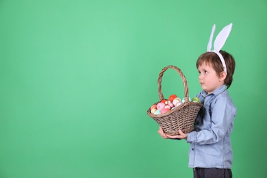 Cute little boy wearing bunny ears with basket full of dyed Easter eggs on green background, space for text