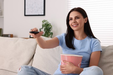 Photo of Happy woman with popcorn bucket changing TV channels with remote control on sofa at home