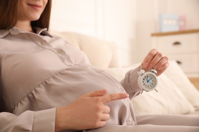 Photo of Young pregnant woman pointing at alarm clock near her belly indoors, closeup. Time to give birth