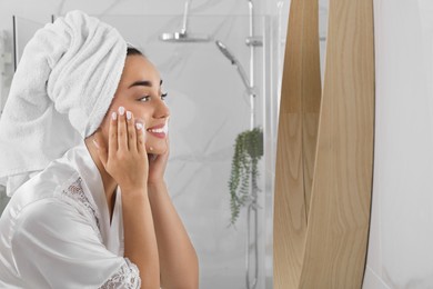 Photo of Beautiful young woman applying cleansing foam onto face near mirror in bathroom, space for text. Skin care cosmetic