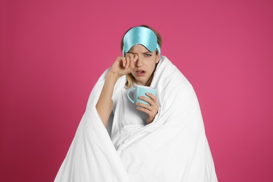 Young woman in sleeping mask wrapped with blanket holding cup on pink background