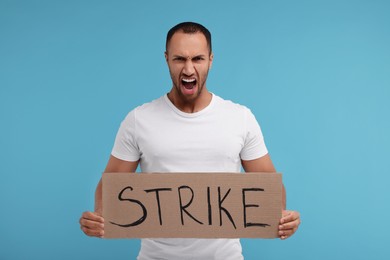 Angry man holding cardboard banner with word Strike on light blue background