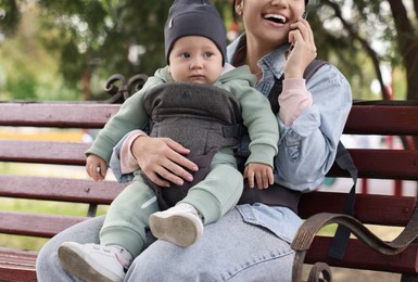Photo of Mother holding her child in sling (baby carrier) while talking on smartphone outdoors, closeup
