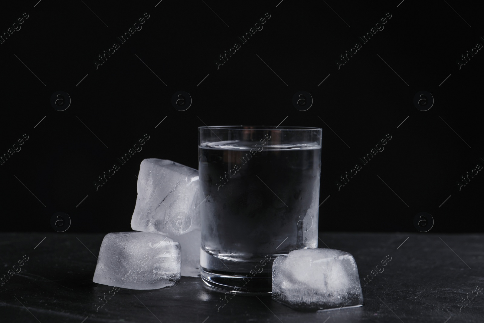 Photo of Vodka in shot glass with ice on table against black background