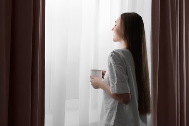 Photo of Woman holding cup of hot drink near window with stylish curtains at home. Space for text