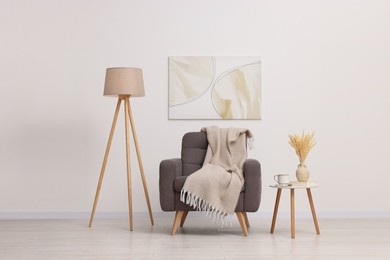 Photo of Comfortable armchair, blanket, lamp and side table near white wall indoors