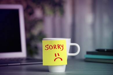 Image of Apology. Sticky note with word Sorry and drawn sad face attached to cup on table in room