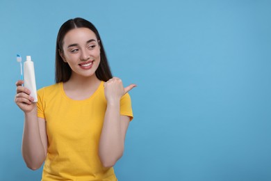 Happy young woman with plastic toothbrush and tube of toothpaste pointing on light blue background, space for text