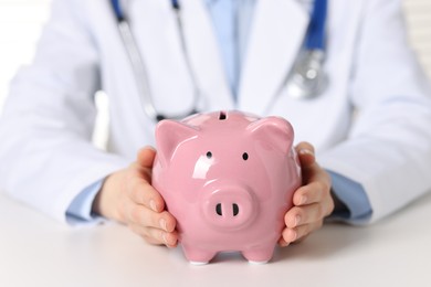 Doctor with piggy bank at white table, closeup