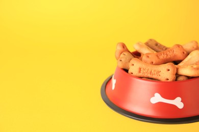 Photo of Bone shaped dog cookies in feeding bowl on yellow background, space for text