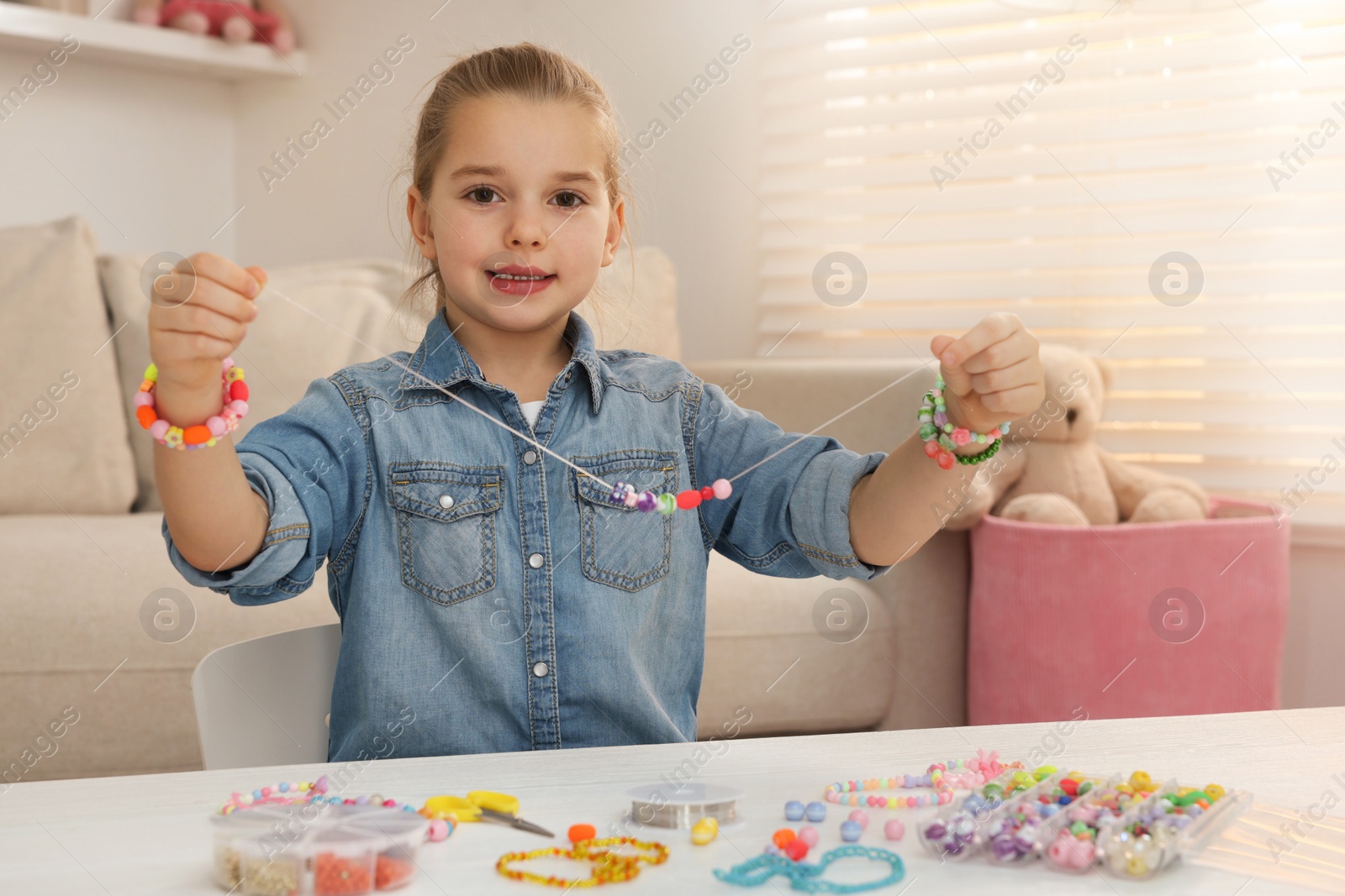 Photo of Cute little girl making beaded jewelry at table in room