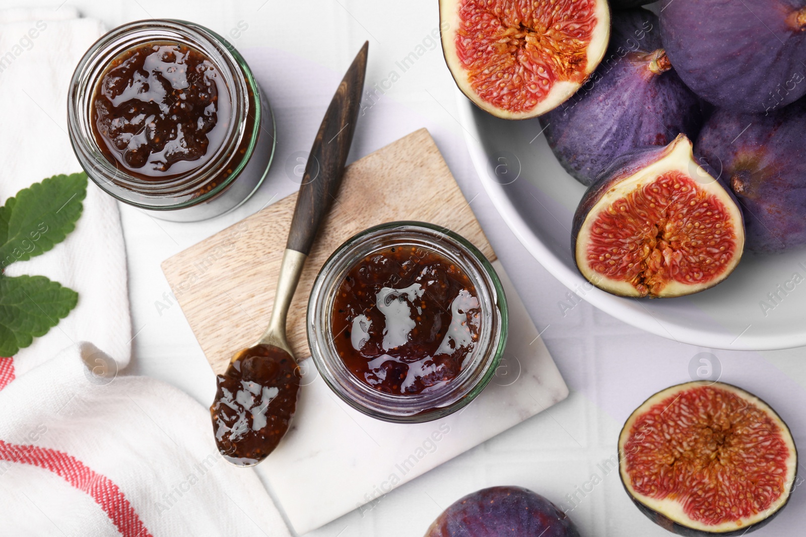 Photo of Glass jars of tasty sweet fig jam and fruits on white tiled table, flat lay