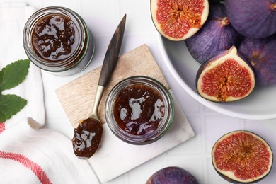 Glass jars of tasty sweet fig jam and fruits on white tiled table, flat lay