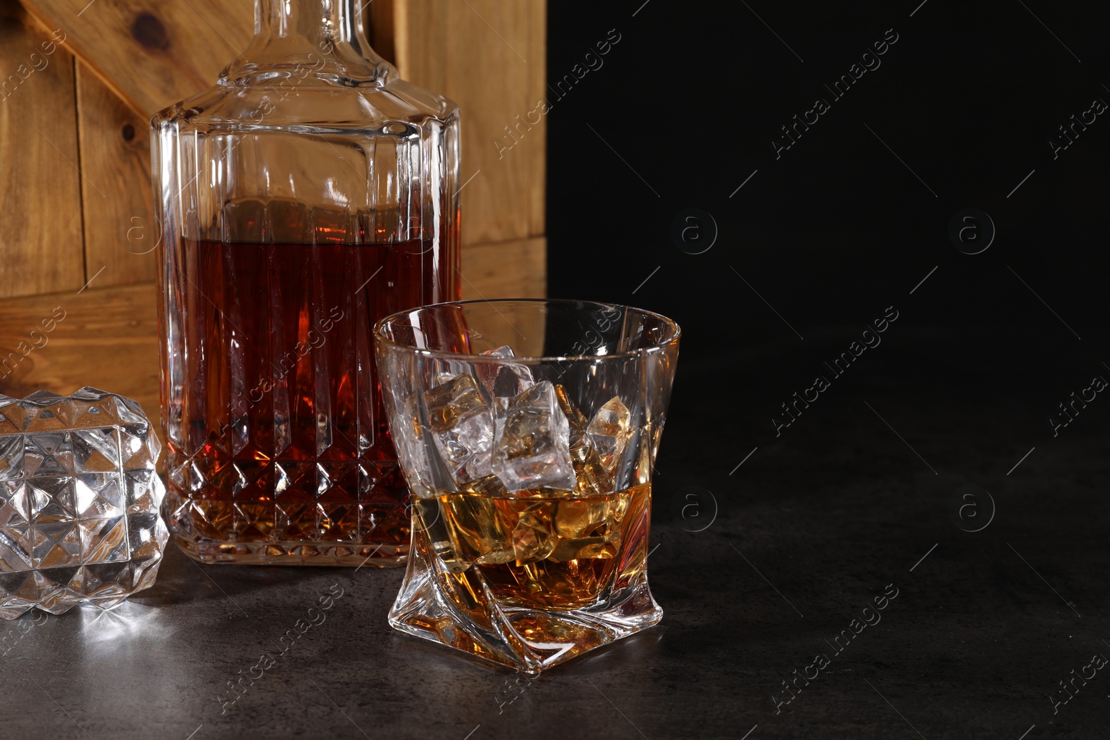 Photo of Whiskey in glass and bottle near wooden crate on dark table against black background. Space for text