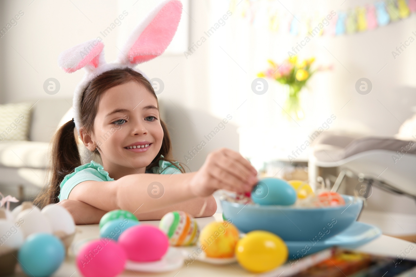 Photo of Cute little girl in bunny ears headband painting Easter eggs at table indoors