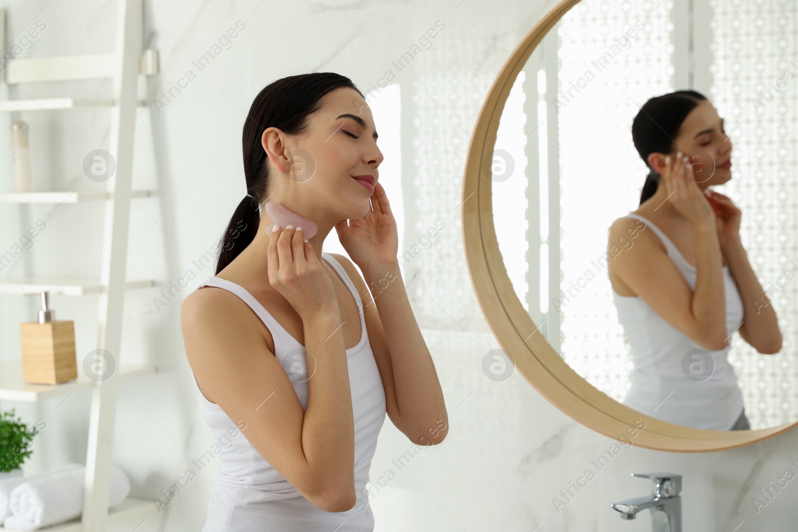 Photo of Beautiful young woman doing facial massage with gua sha tool in bathroom