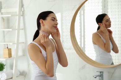 Photo of Beautiful young woman doing facial massage with gua sha tool in bathroom