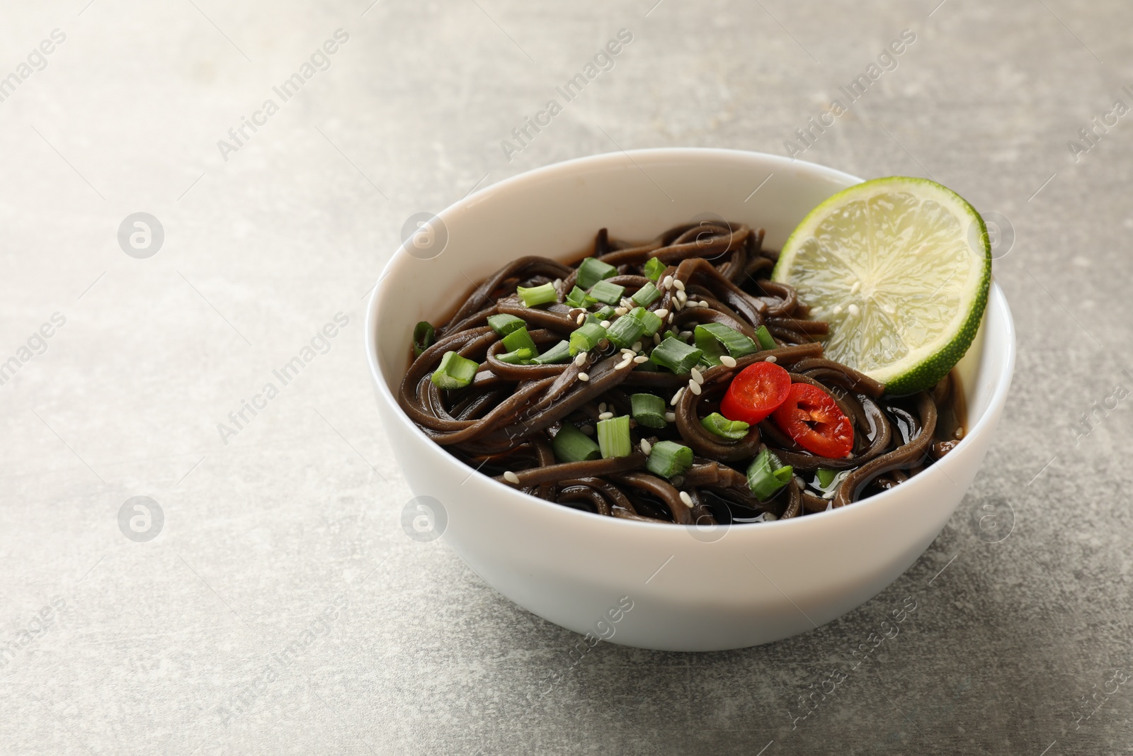 Photo of Tasty buckwheat noodles (soba) with sauce, onion and lime in bowl on light grey table. Space for text