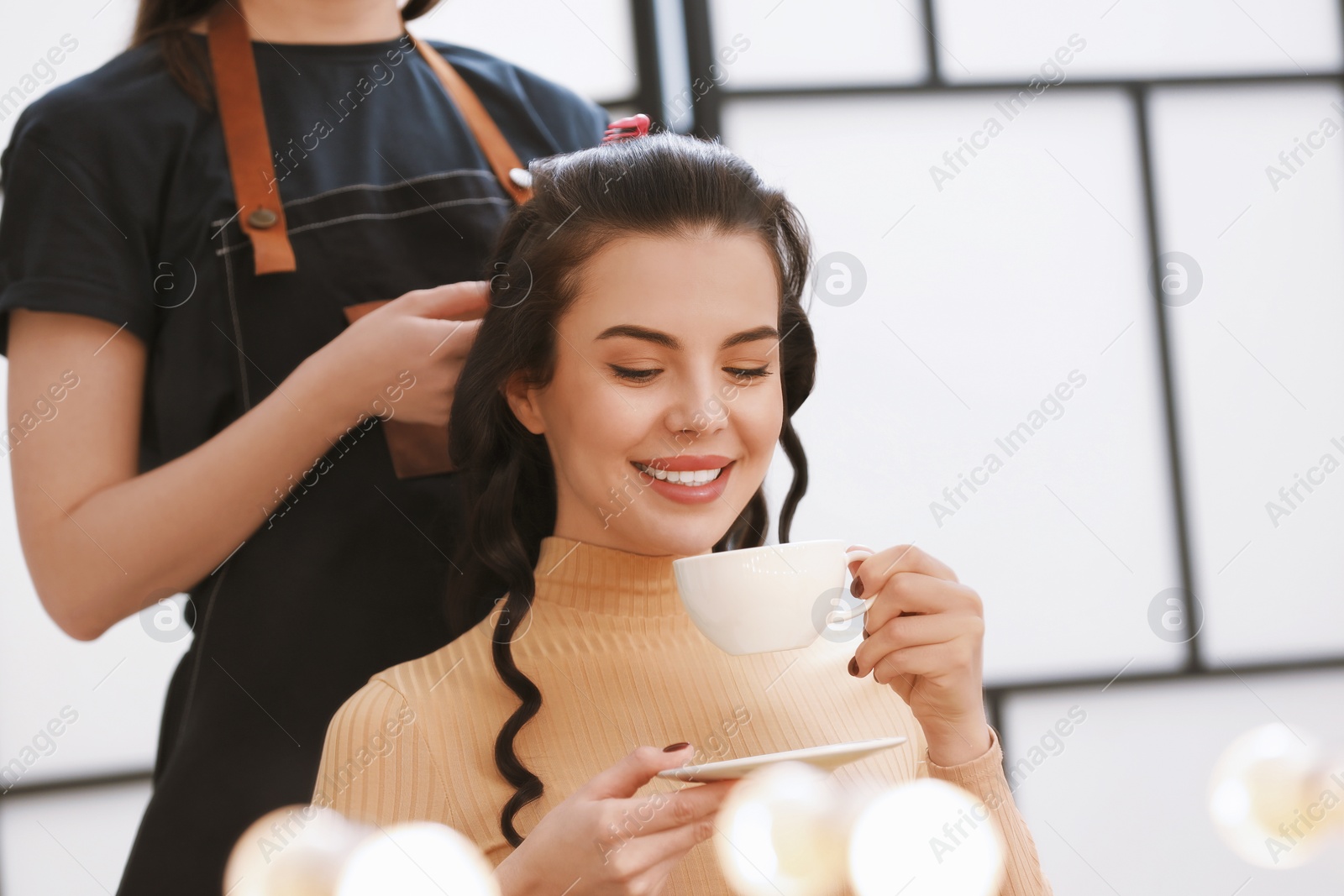 Photo of Hair styling. Professional hairdresser working with smiling woman while she drinking coffee in salon, closeup. Space for text