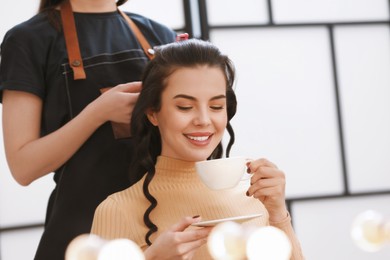 Photo of Hair styling. Professional hairdresser working with smiling woman while she drinking coffee in salon, closeup. Space for text