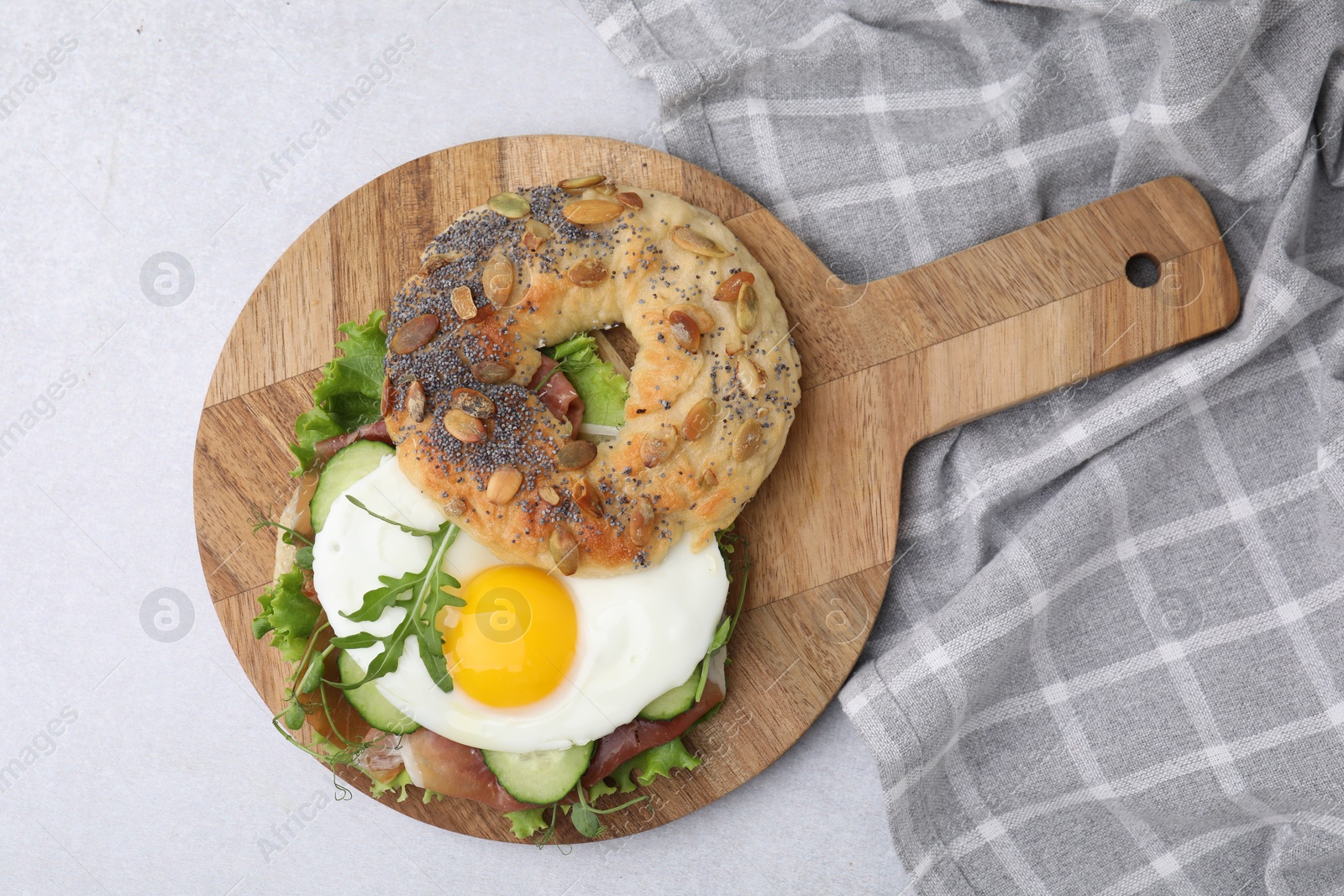 Photo of Tasty bagel with cured ham, egg, cucumber and salad mix on light table, top view