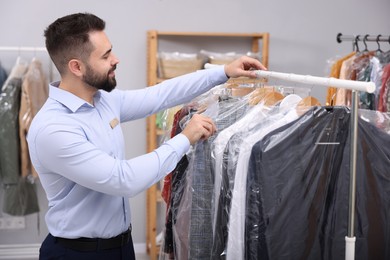 Photo of Dry-cleaning service. Happy worker choosing clothes from rack indoors