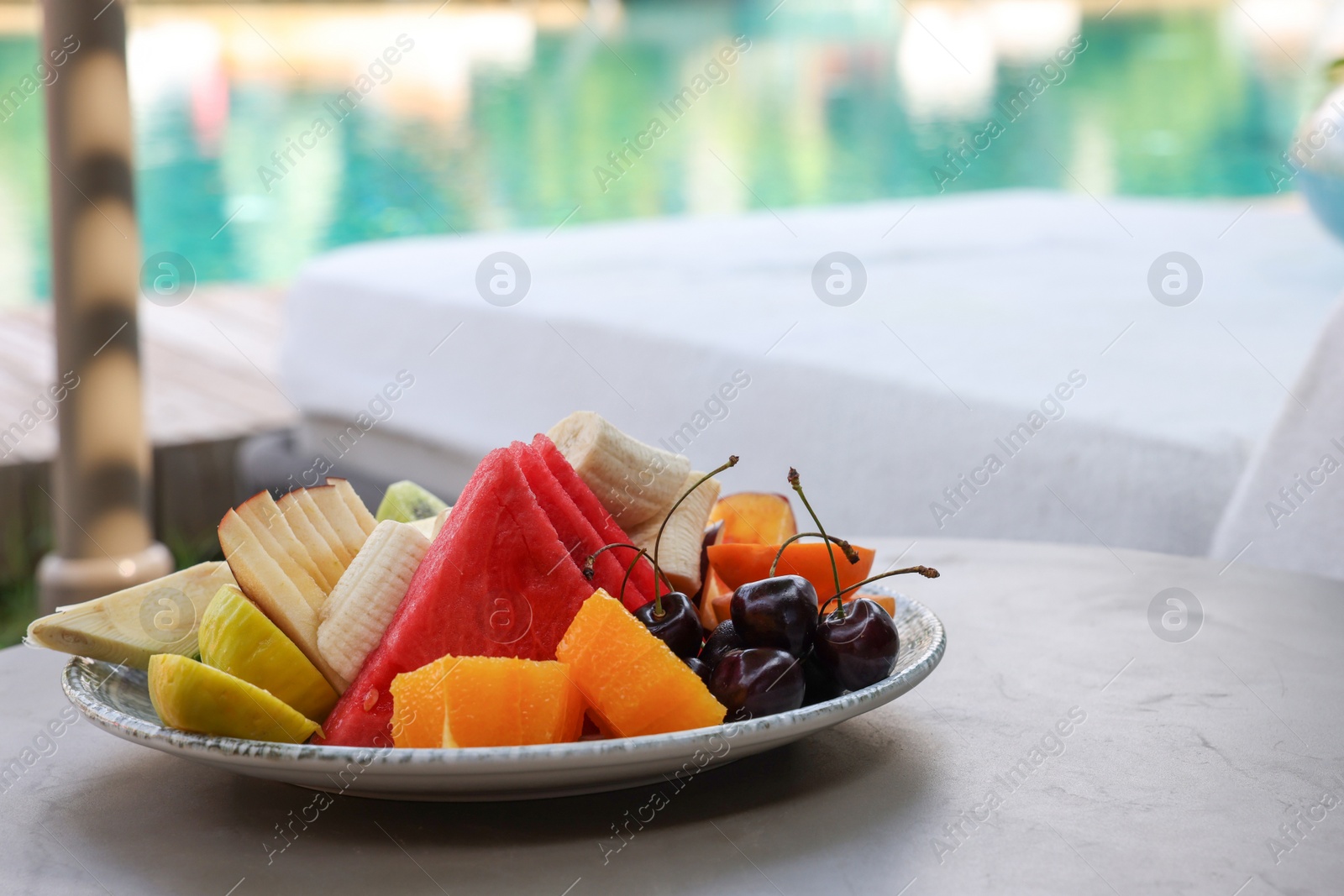 Photo of Plate with fresh fruits on table near sun lounger, space for text. Luxury resort with outdoor swimming pool