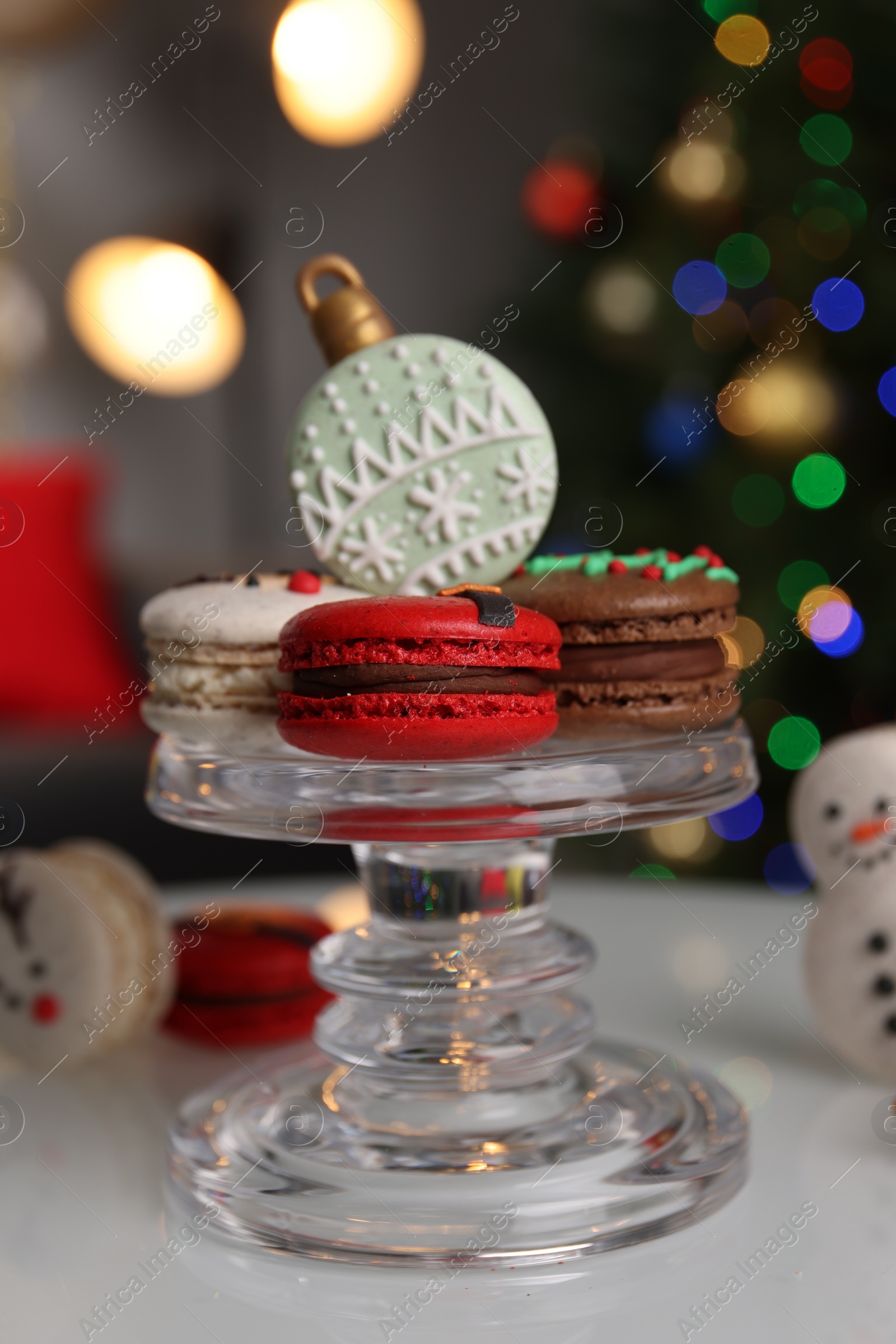 Photo of Beautifully decorated Christmas macarons on white table against blurred festive lights