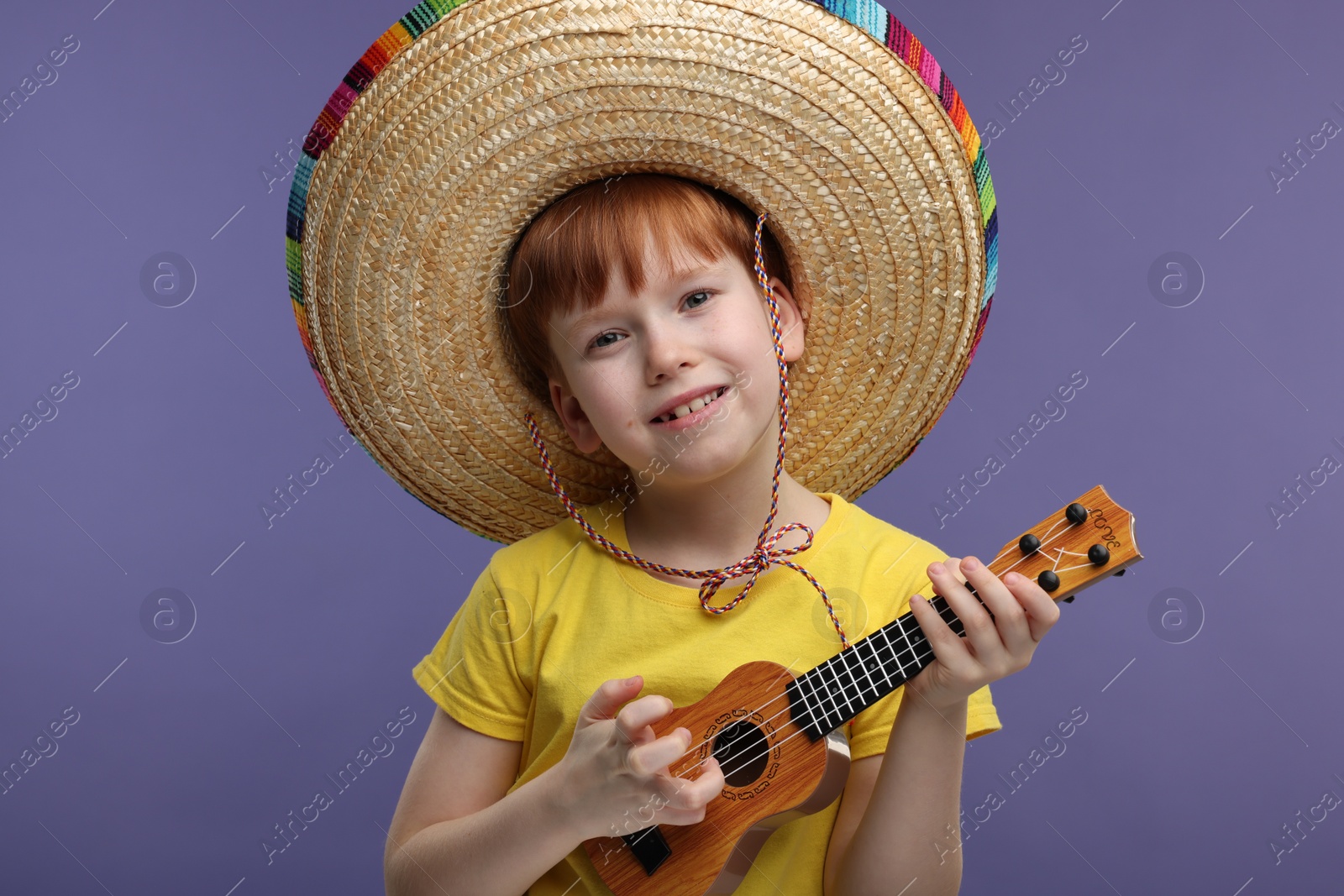 Photo of Cute boy in Mexican sombrero hat playing ukulele on violet background