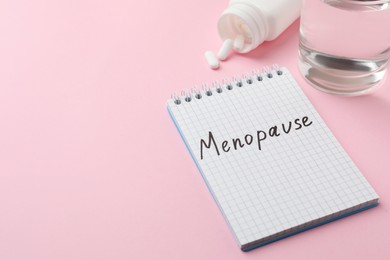 Photo of Notebook with word Menopause near pills and glass of water on pink background, space for text