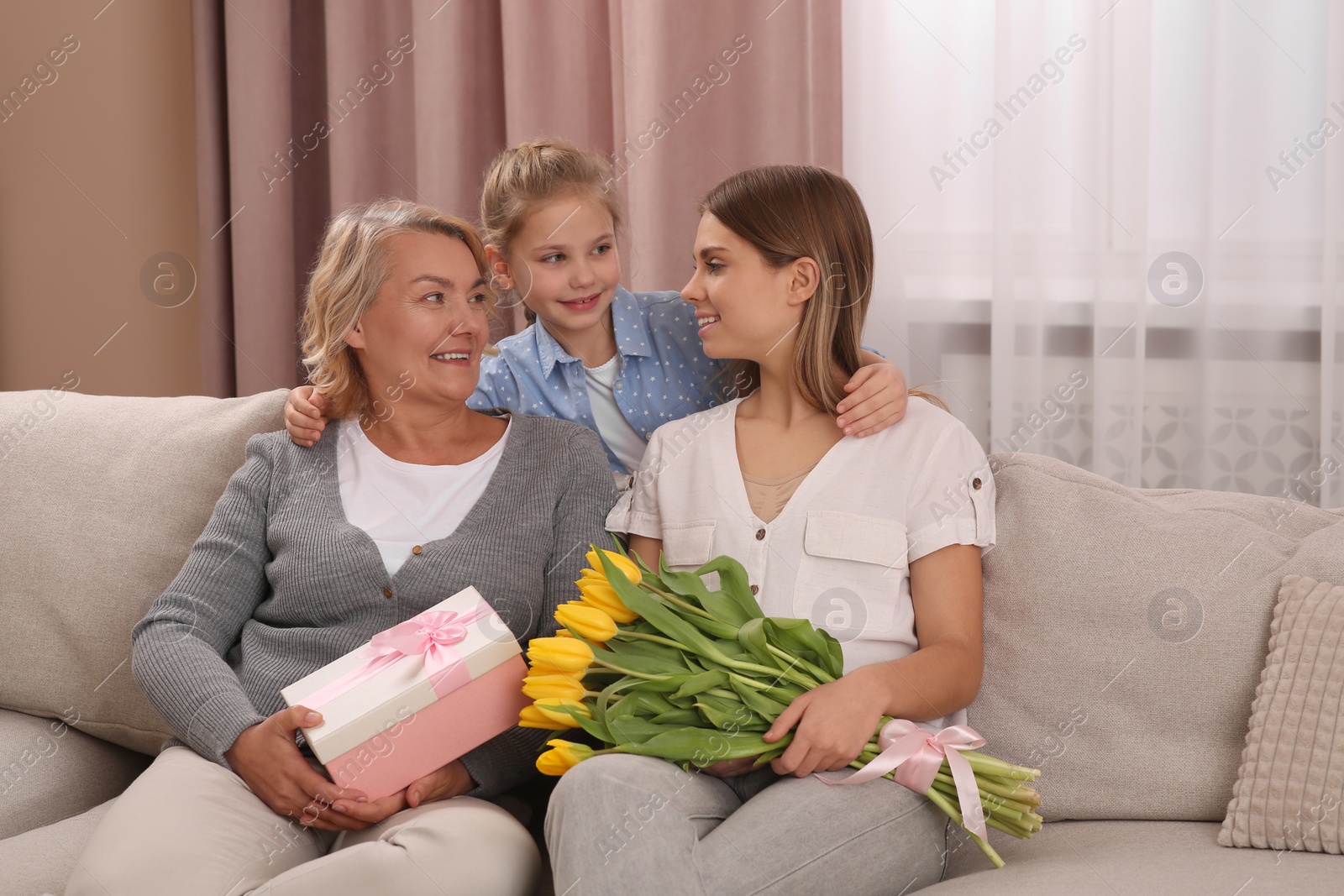 Photo of Little girl congratulating her mom and granny with flowers and gift at home. Happy Mother's Day