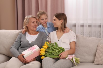 Photo of Little girl congratulating her mom and granny with flowers and gift at home. Happy Mother's Day