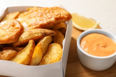 Photo of Disposable container with British Traditional Fish and potato chips on wooden board, closeup