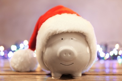 Cute piggy bank with Santa hat on table against blurred Christmas lights