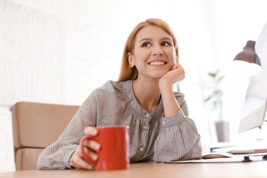 Young woman with cup of drink relaxing at table in office during break