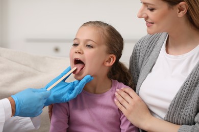 Photo of Doctor examining girl`s oral cavity with tongue depressor near her mother indoors, closeup