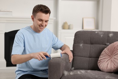Photo of Pet shedding. Smiling man with lint roller removing dog's hair from armchair at home