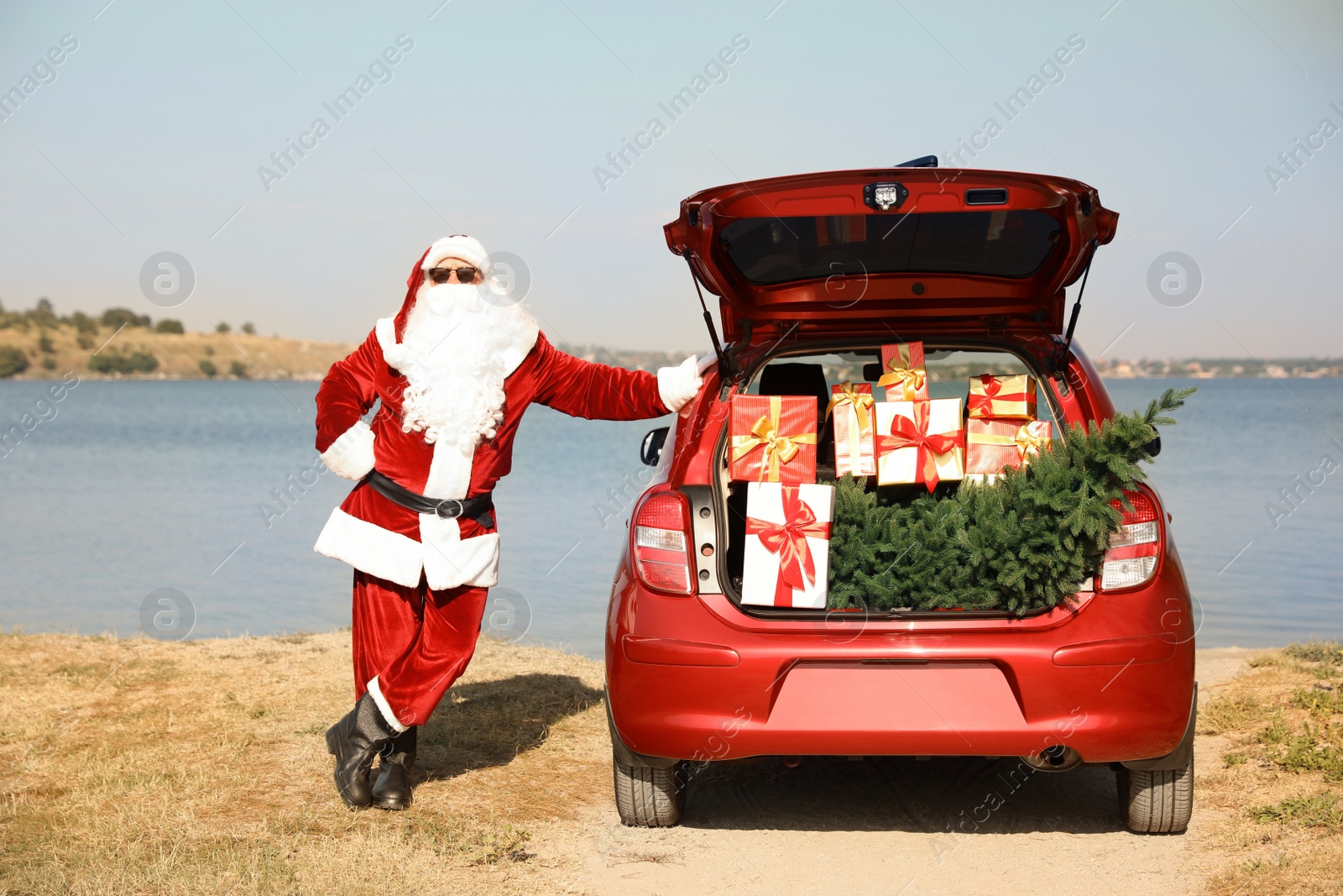 Photo of Authentic Santa Claus near car with open trunk full of presents and fir tree outdoors