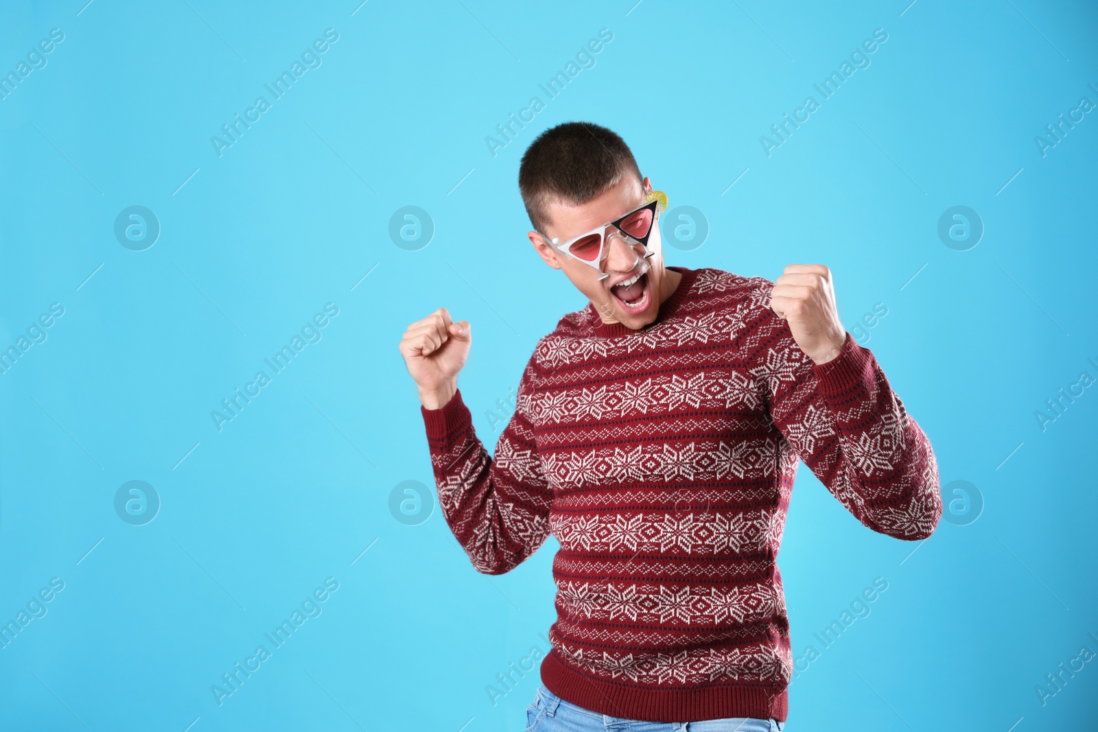 Photo of Emotional young man in Christmas sweater and party glasses on light blue background