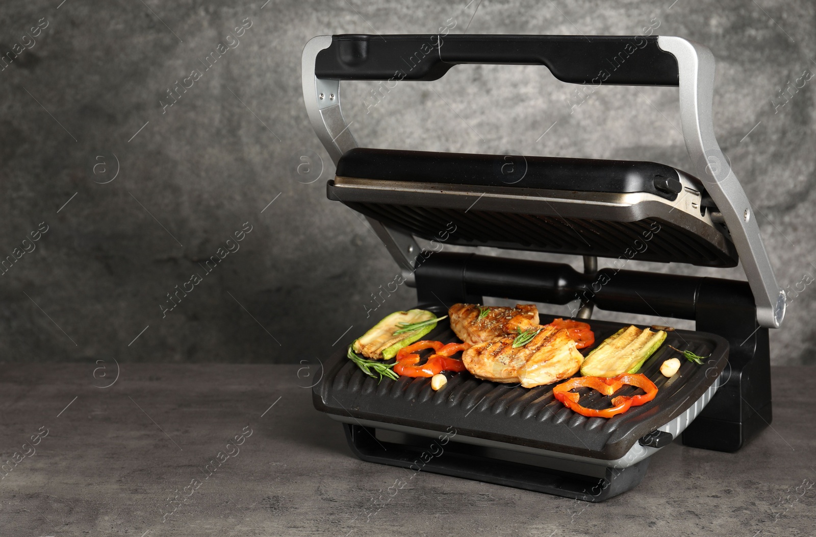 Photo of Electric grill with tasty meat, spices and vegetables on grey textured table, space for text