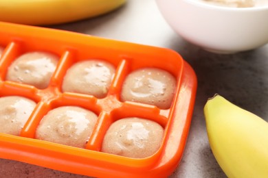 Photo of Banana puree in ice cube tray with fresh banana fruits on grey table, closeup. Ready for freezing