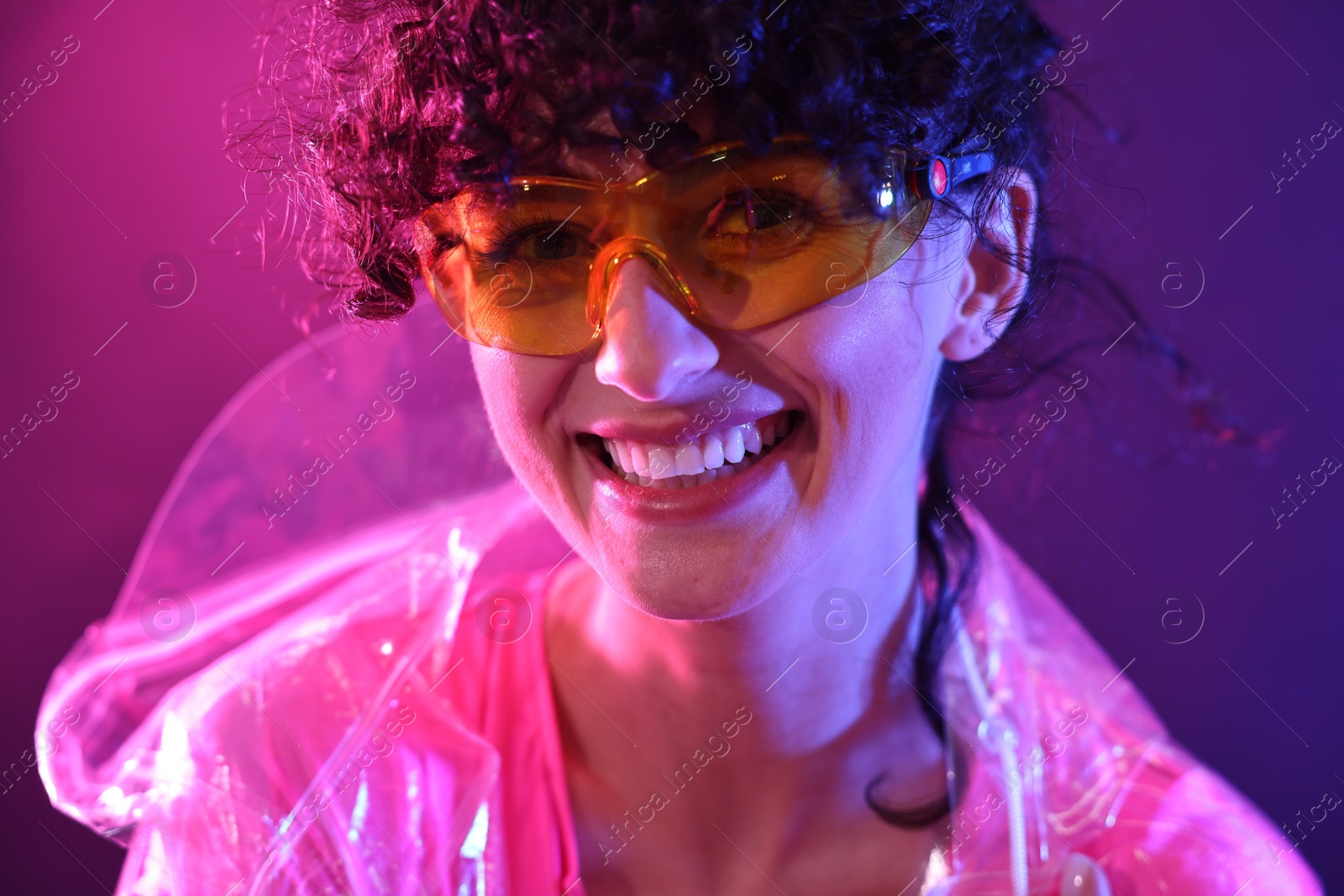 Photo of Beautiful young woman in transparent coat and sunglasses posing on color background in neon lights, closeup