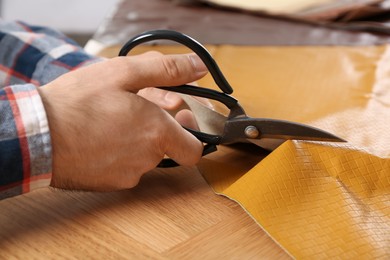 Photo of Man cutting orange leather with scissors at wooden table, closeup