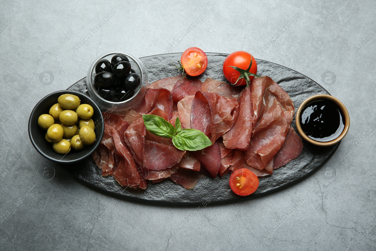 Photo of Delicious bresaola, tomato, olives and basil leaves on grey textured table, top view