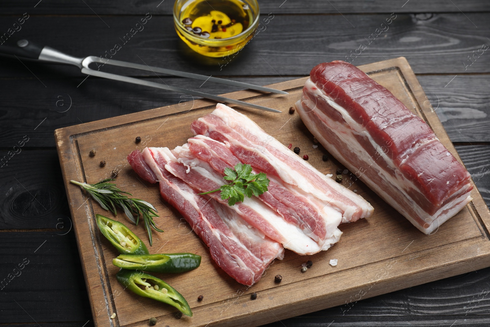 Photo of Pieces of raw pork belly, chili pepper, peppercorns, rosemary and parsley on black wooden table