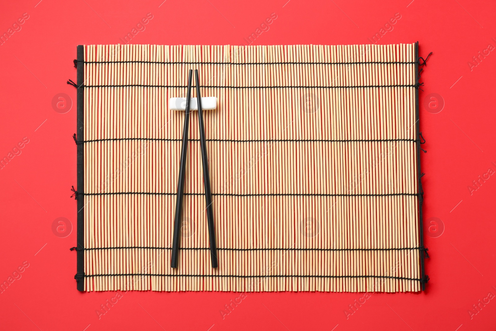 Photo of Bamboo mat with pair of black chopsticks and rest on red background, top view
