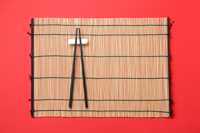 Photo of Bamboo mat with pair of black chopsticks and rest on red background, top view